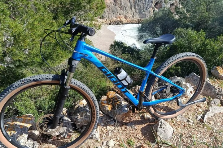 Blue mountain bike parked on rocky terrain with trees and a river in the background.