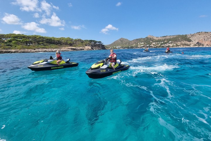 People riding jet skis on clear blue water near a rocky shoreline.