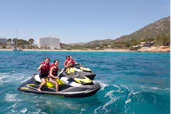 Four people on jet skis in blue water near a beach and hills.