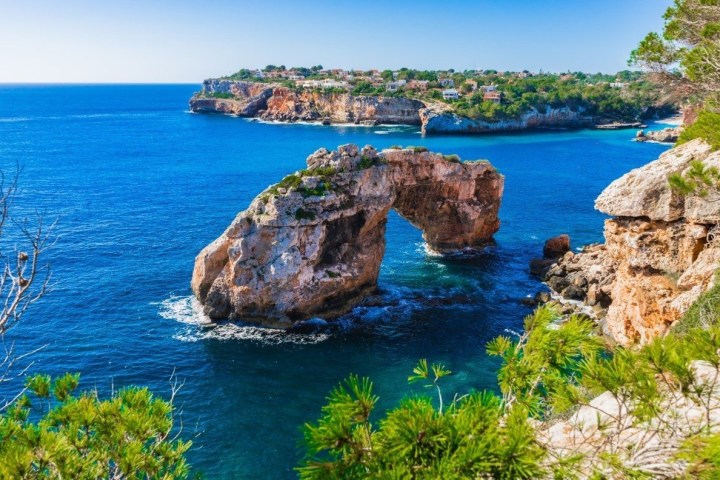 Rock arch in blue sea with rocky coastline and greenery.